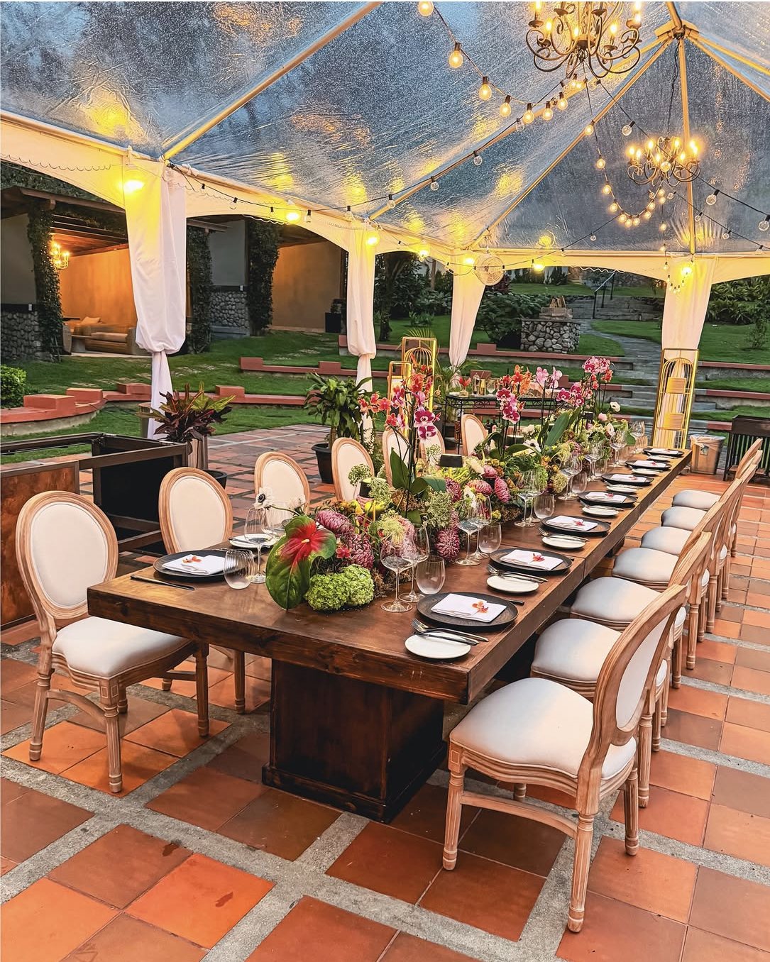 A long wooden table set for dinner with orchids and candles under a clear tent with chandeliers in the hotel garden