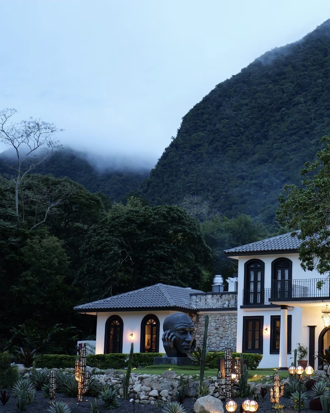 The white villa at dusk with a large bronze head sculpture, glowing garden lamps and the misty mountain behind