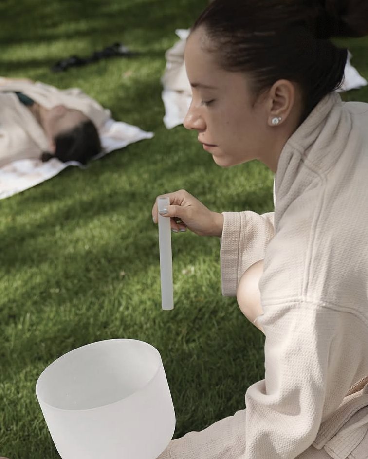 A practitioner holding a crystal singing bowl on the lawn while women rest on blankets in the grass